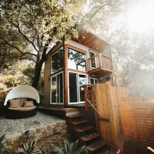 Cabane moderne en bois avec grandes baies vitrées, entourée d’arbres, avec un coin détente extérieur.