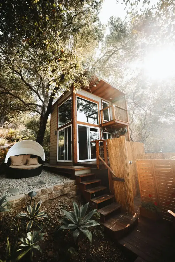 Cabane moderne en bois avec grandes baies vitrées, entourée d’arbres, avec un coin détente extérieur.