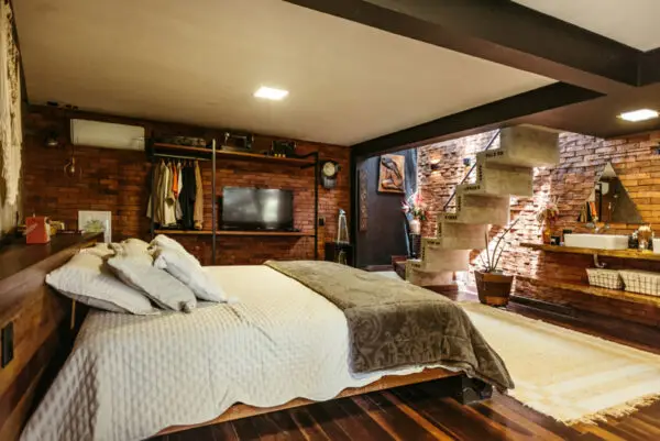 Chambre chaleureuse au design industriel avec murs en briques, lit double, escalier en béton et éclairage doux.